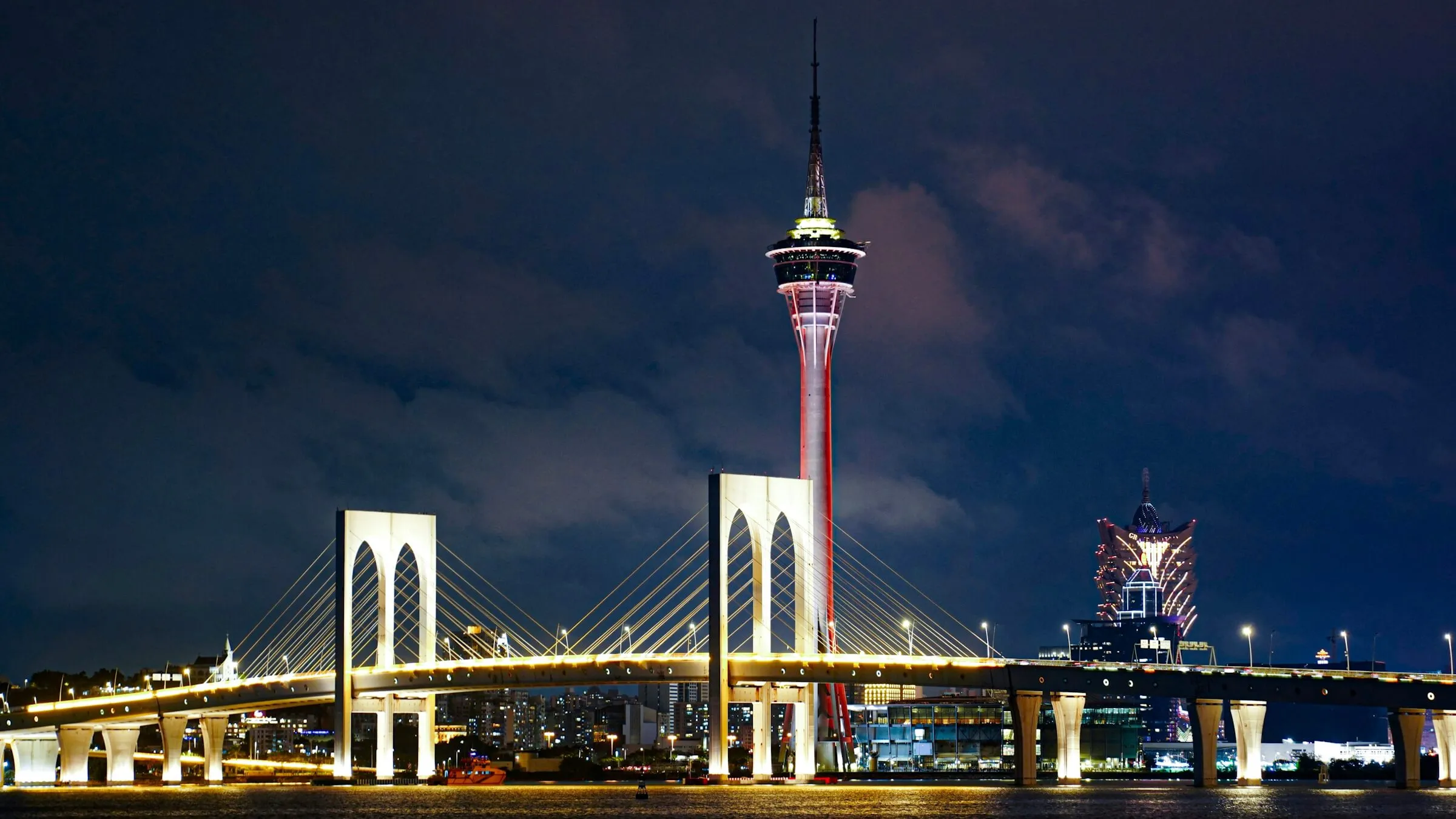 Macau Tower lit up red and white at night, seen across the water behind the illuminated Sai Van Bridge, with the Grand Lisboa on the right
